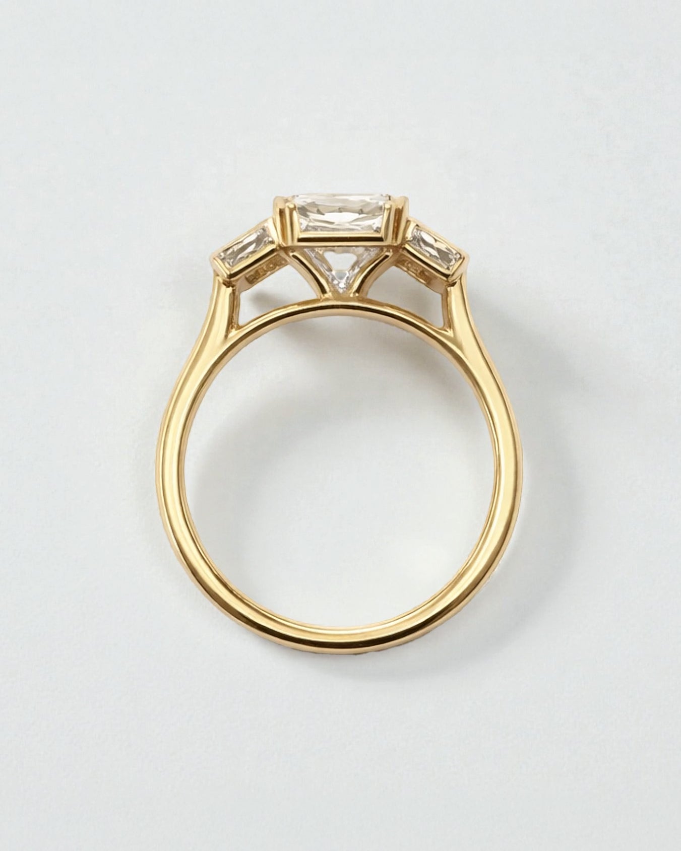 Angled view of 3ct emerald cut lab grown diamond bezel trilogy ring with baguette side diamonds in yellow gold