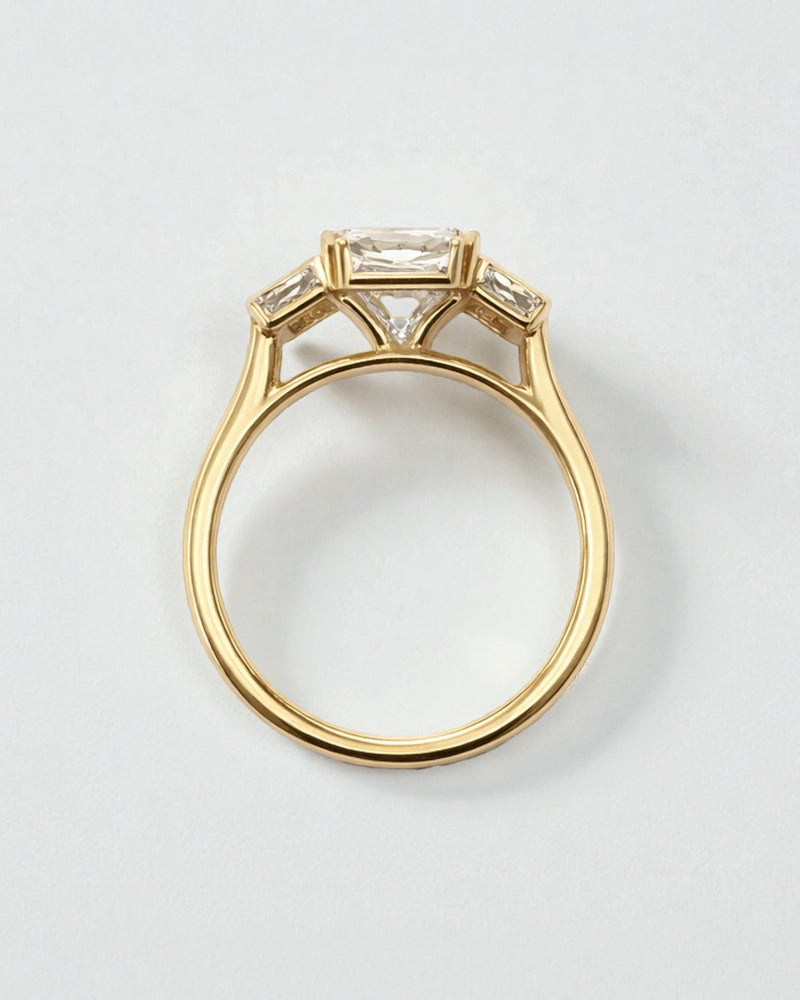 Angled view of 3ct emerald cut lab grown diamond bezel trilogy ring with baguette side diamonds in yellow gold